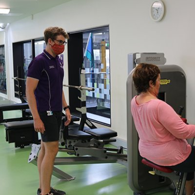Woman using lat pulldown machine under supervision of man in UQ shirt